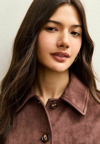 Chaqueta de ante marrón con un cuello con muesca, dos botones y una textura suave. El cabello es largo y ondulado, lo que pone el foco en la prenda.