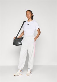 Kvinne i hvit Adidas t-skjorte, hvite joggebukser med rosa striper og sneakers, som holder en svart Adidas treningsbag med skulderreim.