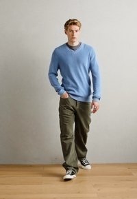 Błękitny sweter w serek na szarym T-shircie, w połączeniu z oliwkowymi spodniami i czarno-białymi sneakersami, stojąc na drewnianej podłodze.