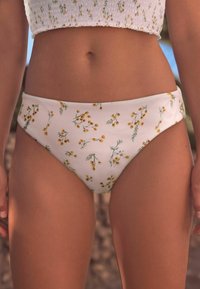 Bílé bikiny s květinovým vzorem, na kterém jsou žluté květy a zelené listy. Měkký materiál, střední výška a hladká textura.
