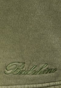 Tissu tricoté vert avec texte cursif brodé en vert olive près du bord inférieur, montrant une texture de fil détaillée.