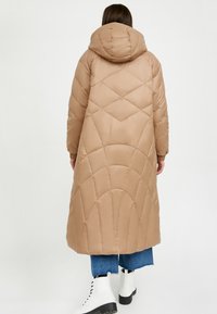 Lange, beige pufferjas met een capuchon en een diamantgequilt design. Heeft geribbelde manchetten en valt tot halverwege de kuit, gecombineerd met blauwe jeans en witte laarzen.