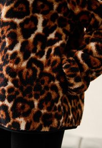 Pull en polaire imprimé léopard avec une texture douce, des couleurs chaudes marron et noir, et un design décontracté avec une poche latérale.