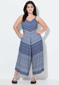 Salopette a righe in blu navy e bianco, con scollatura a V e design a gamba larga. Presenta spalline sottili e un tessuto textured. Indossata con sandali argentati.