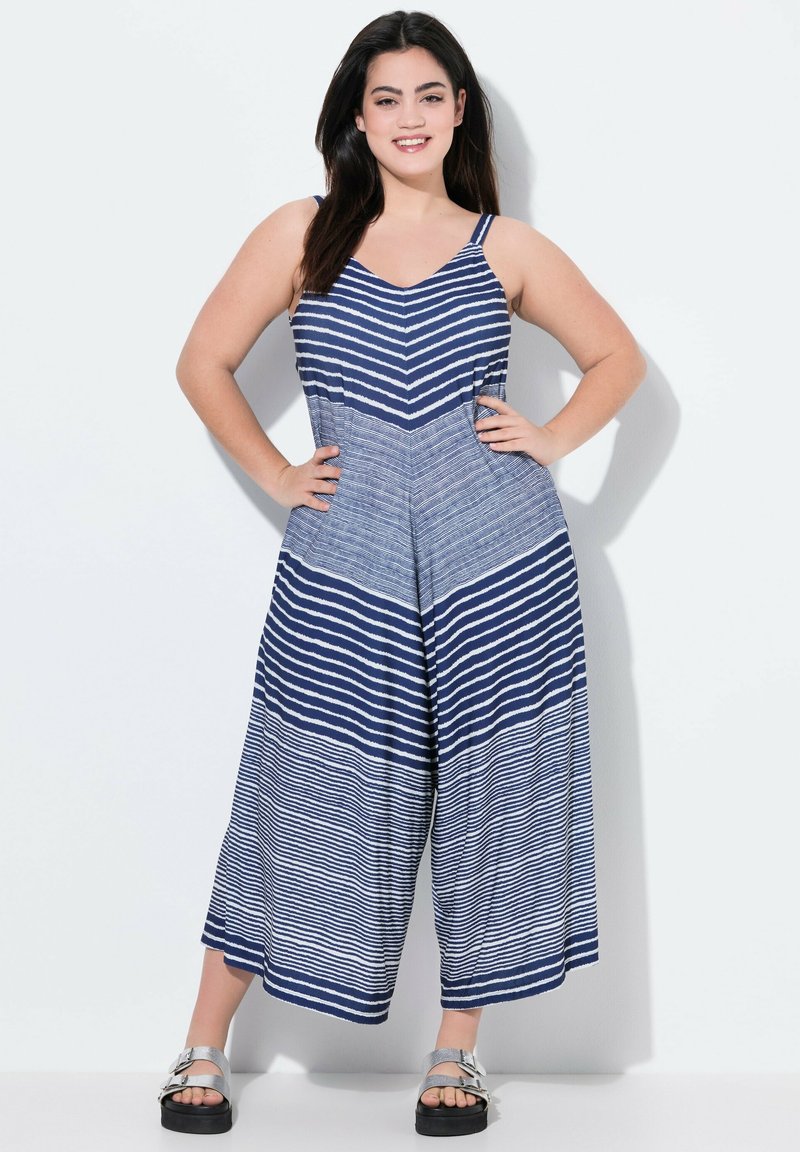 Salopette a righe in blu navy e bianco, con scollatura a V e design a gamba larga. Presenta spalline sottili e un tessuto textured. Indossata con sandali argentati.