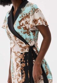 Robe portefeuille florale en menthe, crème et noir. Dotée de manches courtes, d'un col noir et d'une taille nouée. Le motif comprend des détails en or complexes.