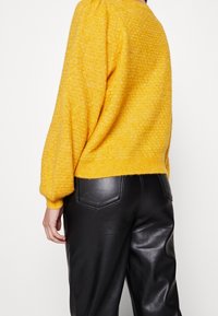 Personne portant un pull jaune texturé avec des manches bouffantes et un pantalon en cuir noir, vue de dos sur un fond blanc.
