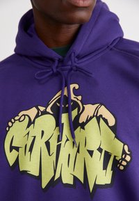 Paarse hoodie met een grafisch ontwerp dat groene tekst en een karakterillustratie bevat. De materialen lijken zacht met een capuchon met trekkoord en een kangoeroezak.