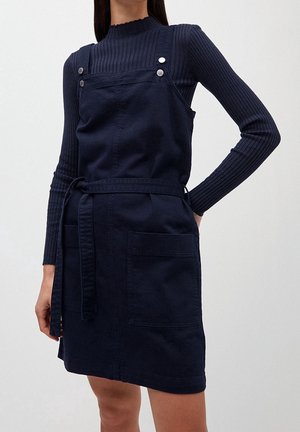 Nainen pukeutuneena laivastonsiniseen essumekkoon, jossa on etutaskut, päällään ribbineuloksinen pitkähihaisen laivastonsininen pooloneule.