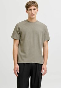 Noor mees lühikeste heledate pruunide juustega, kes kannab tekstuuriga oliivrohelist lühikeste varrukatega särki ja musti pükse, seisab lihtsa valge tausta ees.