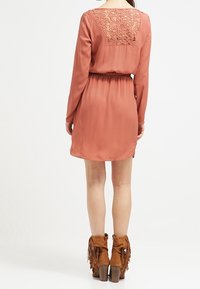 Vestido de color óxido con mangas largas y detalle de encaje en la espalda; cinturilla elástica, textura suave, combinado con botas de tobillo marrones con flecos.