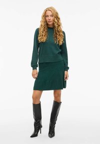 Pulover verde tricotat cu mâneci lungi, combinat cu o fustă asortată până la genunchi. Purtați cu cizme negre din piele, elegante și cu toc înalt.