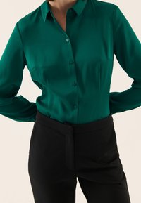 Camicia in seta teal con bottoni, colletto e maniche lunghe, abbinata a pantaloni neri sartoriali con vita e design elegante.