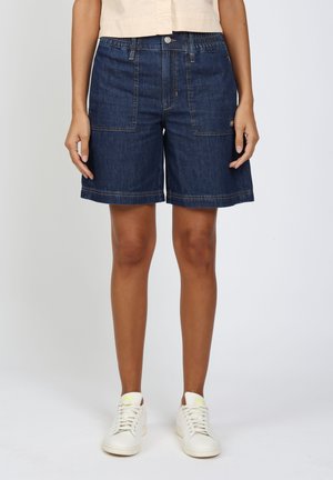 Person trägt dunkelblaue Jeansshorts mittlerer Länge mit aufgesetzten Fronttaschen und weiße Sneakers, steht vor einem einfarbigen Hintergrund.