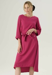 Abito fucsia con maniche lunghe, scollatura rotonda, cintura e orlo asimmetrico. Presenta una texture liscia e sottili dettagli con bottoni.