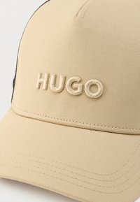 Μπεζ καπέλο μπέιζμπολ από υφασμάτινη υφή, με κεντημένο λογότυπο "HUGO" σε ανοιχτό μπεζ χρώμα. Διαθέτει καμπυλωτή γείσο και ραμμένες λεπτομέρειες.