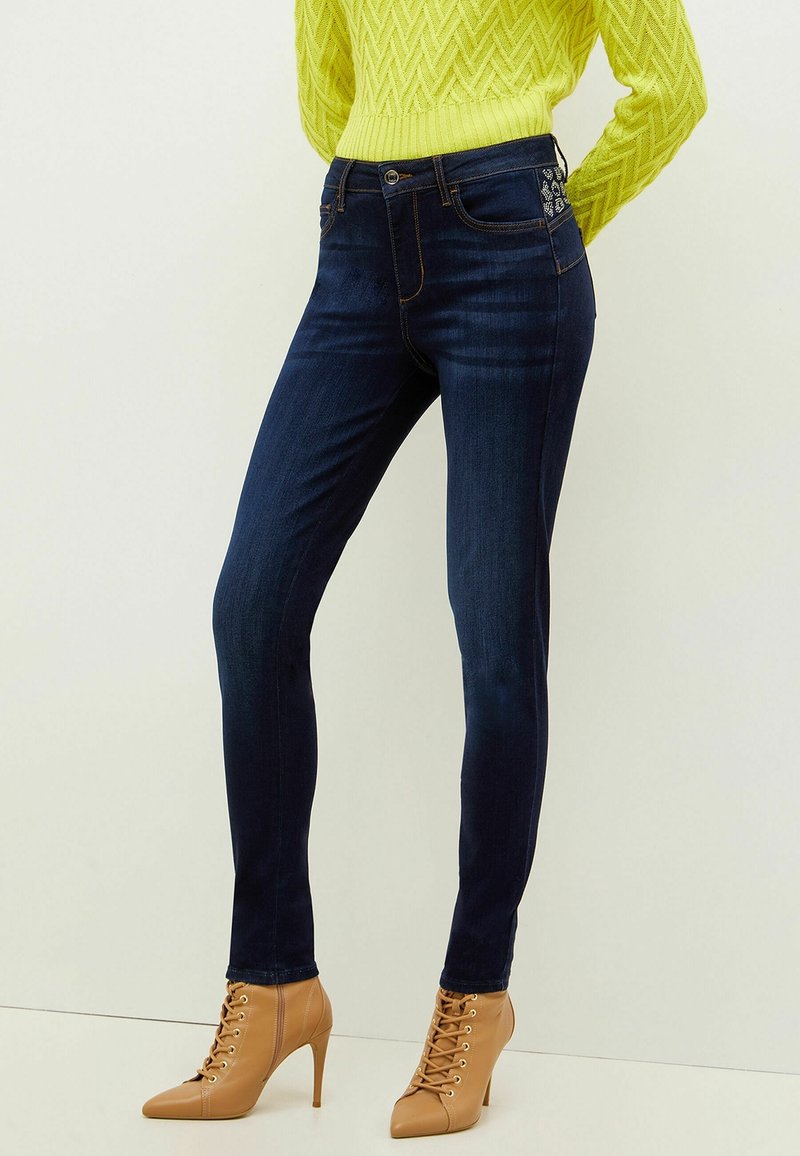 Kvinna som bär mörkblå skinny jeans med broderade fickdetaljer, gul kabelstickad tröja och bruna högklackade snörstövlar.