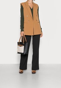 Braun doppelt knöpfbare Weste über einem dunkelgrünen Pullover, kombiniert mit schwarzen Hosen und einer schwarzen Handtasche mit cremefarbenem Akzent. Braune Absatzstiefel.