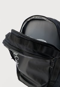 Μαύρο σακίδιο πλάτης με το κύριο διαμέρισμα ανοιχτό, που δείχνει το άδειο εσωτερικό και ένα μερικώς ορατό λευκό λογότυπο στην μπροστινή τσέπη.