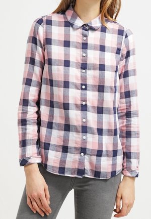 Kobieta ubrana w zapinaną na guziki koszulę z długim rękawem o różowo-biało-granatowym wzorze w kratę, zestawioną z szarymi spodniami.