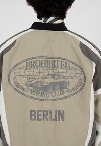 Personne portant une veste beige et grise avec le texte "Prohibited Racing Club" et un graphisme de voiture, ainsi que le texte "Berlin" dans le dos.
