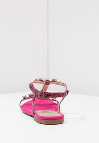 Růžové ploché sandály s hadím vzorem, zdobené zlatými a modrými drahokamy na bílém povrchu a pozadí.
