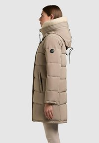 Donna in profilo che indossa un piumino trapuntato beige con colletto alto foderato in pile e cappuccio.