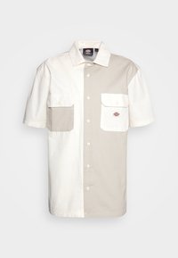 Camicia a maniche corte con design diviso in beige e crema, presenta due tasche frontali e bottoni bianchi, realizzata in tessuto di cotone.
