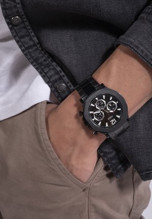 Montre chronographe noire avec des accents argentés, cadran octogonal et trois sous-cadrans. Bracelet en métal, porté au poignet, associé à une chemise grise et un pantalon beige.