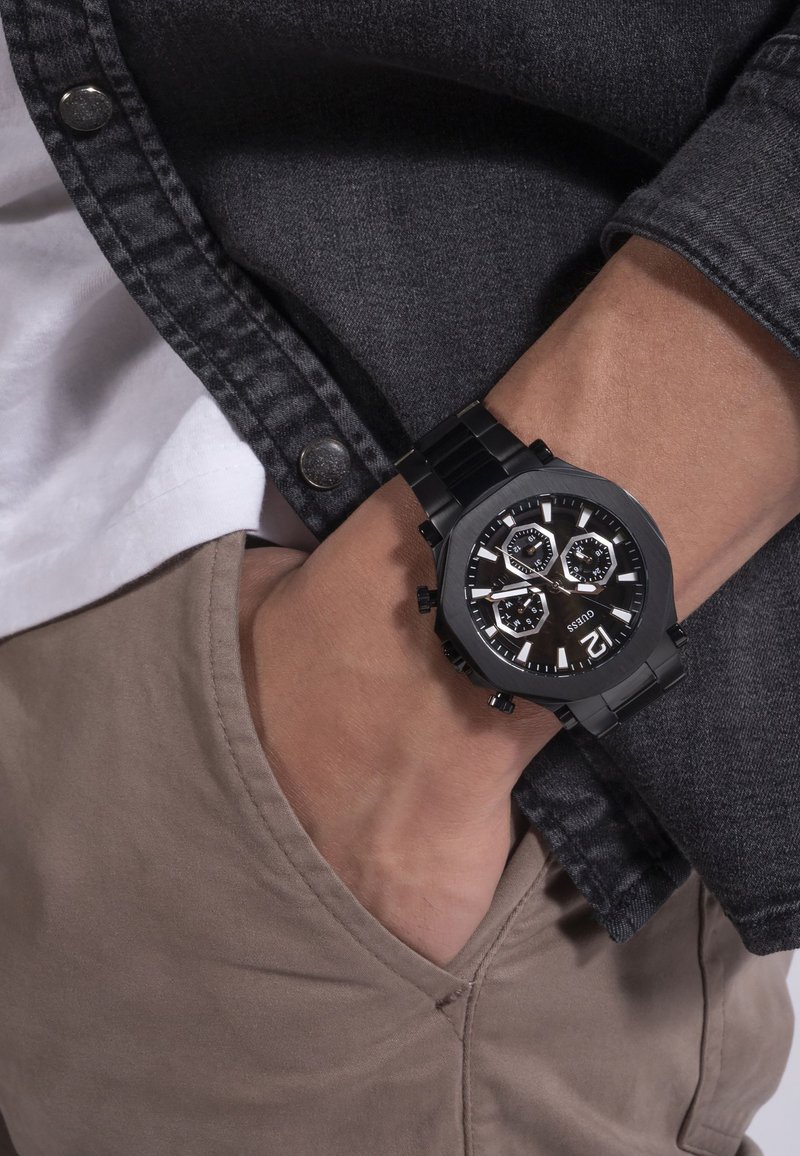 Czarny analogowy zegarek z okrągłą tarczą, trzema podtarcze, żłobioną obręczą i bransoletą ze stali nierdzewnej. Noszony na nadgarstku z widoczną tkaniną w odcieniu karmelowym.