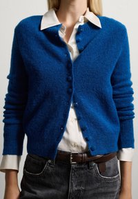 Blauwe gebreide cardigan met een zachte textuur, voorzien van een knoopsluiting en ribgebreide manchetten, gedragen over een witte blouse met kraag en donkere jeans.