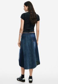Falda de mezclilla con diseño acampanado, en diferentes tonos de azul y con bolsillos traseros. Combinada con una blusa corta negra y zapatillas negras.