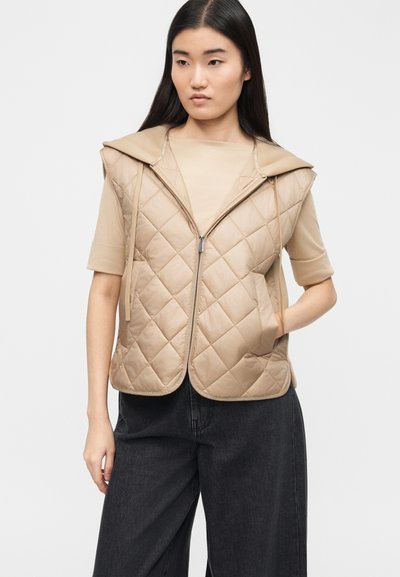 Νέα γυναίκα που φοράει ένα μπεζ γιλέκο με κουκούλα χωρίς μανίκια, ραμμένο σε σχέδιο, πάνω από ένα μπεζ κοντομάνικο πουκάμισο και μαύρο παντελόνι με φαρδιά μπατζάκια, στέκεται με το ένα χέρι στην τσέπη.