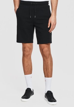 Mężczyzna ubrany w czarne szorty z ściągaczem, białe skarpetki sportowe i czarne tenisówki, stojący na jednolitym jasnym tle.