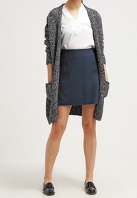 Cardigan lungo grigio a maglia con apertura frontale e tasche, indossato sopra una blusa bianca e una gonna blu scuro, abbinato a mocassini neri.