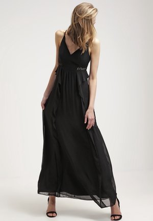 Femme portant une robe longue noire fluide sans manches avec des détails en dentelle et des volants, debout devant un fond clair uni.