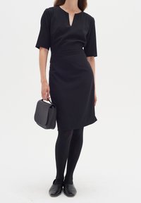 Svart klänning med korta ärmar, v-formad halsringning, figursydd midja, knälång kjol, matchad med svarta strumpbyxor och skor, håller en svart handväska.