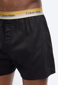 Μαύρα μποξεράκια Calvin Klein με ελαστική μέση με κίτρινο διαβαθμισμένο χρώμα, φορεμένα από ένα άτομο με μέτριου-σκούρου τόνου δέρμα.