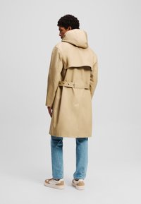 Trench beige con capucha, cinturón en la cintura y detalle en la parte trasera estilo capa. El modelo lleva puestos unos jeans azul claro y zapatos marrones.