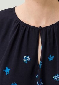 Blouse bleu marine avec un col froncé, une patte de boutonnage avec un bouton rond et une petite broderie florale en bleu et vert.