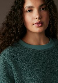 Mørk teal strikket sweater med rund halsudskæring og ribbet kant. Har en blød, fleece-lignende overflade og en afslappet pasform.