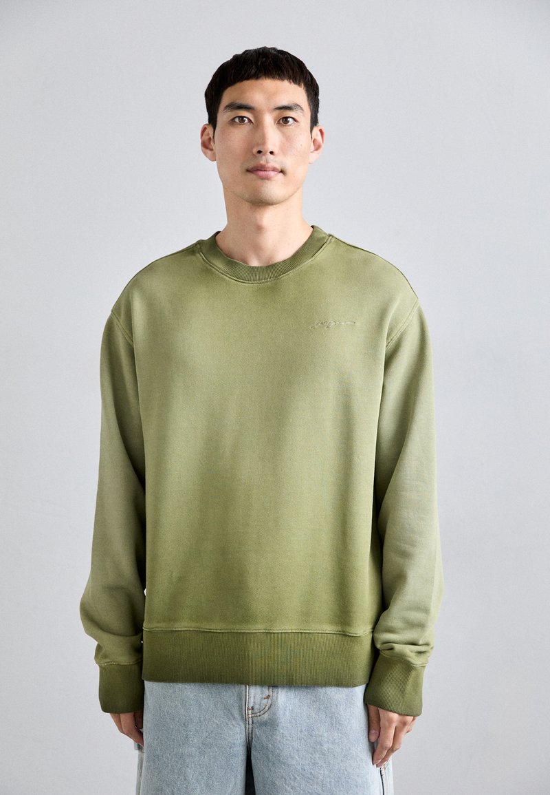 Felpa verde oliva con colletto e polsini a coste, caratterizzata da una vestibilità rilassata e un branding discreto sul petto. Indossata con jeans azzurri chiari.