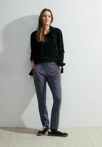 Grå joggerbyxor med dragsko i midjan, matchat med en svart topp och en svart texturerad jacka, avslutat med svartvita sneakers.