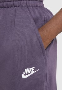 Lila melegítőnadrág, rugalmas derékkal, fehér hímzett Nike logóval és oldalsó zsebbel, puha, texturált anyagból.
