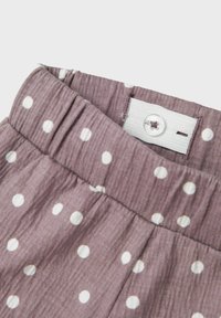 Tissu violet avec des pois blancs, doté d'une taille froncée et d'une fermeture à bouton. Texture douce, avec des détails de couture visibles.