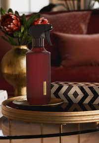 Rødt glassprayflaske med rillet tekstur, svart avtrekkerdyse og gulletikett på et dekorativt brett, omgitt av mønstret dekor.