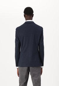 Sötétkék gyapjú blézer slim fit szabásban, iker hátsó szellőzőkkel, gombos mandzsettákkal, szürke nadrágokkal párosítva. Sima textúra.