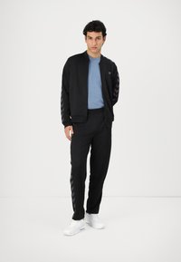 Zwarte sportieve trainingsoutfit met een ritsjack met chevron-gepatenteerde mouwen, gecombineerd met zwarte broeken en aangevuld met witte sneakers.
