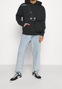 Sudadera negra con bolsillo frontal y acentos de logo en blanco. Combinada con jeans rectos de un azul claro y zapatillas negras con detalles en blanco.