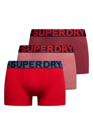 Trzy męskie bokserki w jednolitym czerwonym, czerwonym melanżowym i ciemnoczerwonym kolorze, każde z szerokim pasem z logo „SUPERDRY” w kontrastowych kolorach.